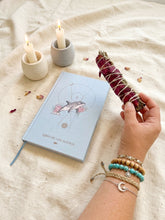 Cargar imagen en el visor de la galería, El Sahumerio Lefay, elaborado con salvia blanca y pétalos de rosa secos, es un sahumerio artesanal diseñado para rituales de limpieza energética, meditación y bienestar. En la imagen aparece sostenido sobre un altar con velas encendidas y el Libro de los Sueños, creando un ambiente de calma, intención y conexión espiritual.