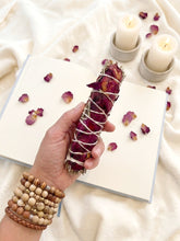 Cargar imagen en el visor de la galería, Primer plano del Sahumerio Lefay hecho a mano con salvia blanca natural y rosas secas, utilizado para limpiar la energía de personas y espacios. El sahumerio se presenta sobre el libro de los suenos, velas y pétalos de rosa, evocando un ritual de autocuidado, mindfulness y armonización del hogar.